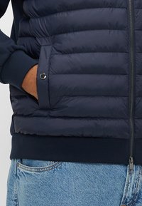 Laivastonsininen toppatakki, jossa vaakasuuntainen tikattu kuvio, vetoketjusuljenta, resorit hupun ja hihansuiden kohdalla sekä sivutasku, joka on korostettu hopeisella yksityiskohdalla.