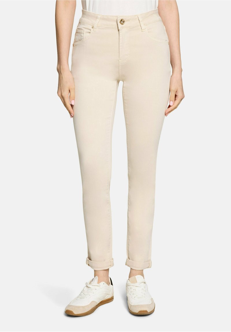 Femme portant un pantalon beige slim retroussé et des baskets blanches, debout devant un fond blanc uni.