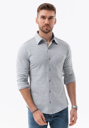 Camisa gris de manga larga con cuello abotonado, que presenta una textura suave de mezcla de algodón y mangas enrolladas, combinada con jeans azules.