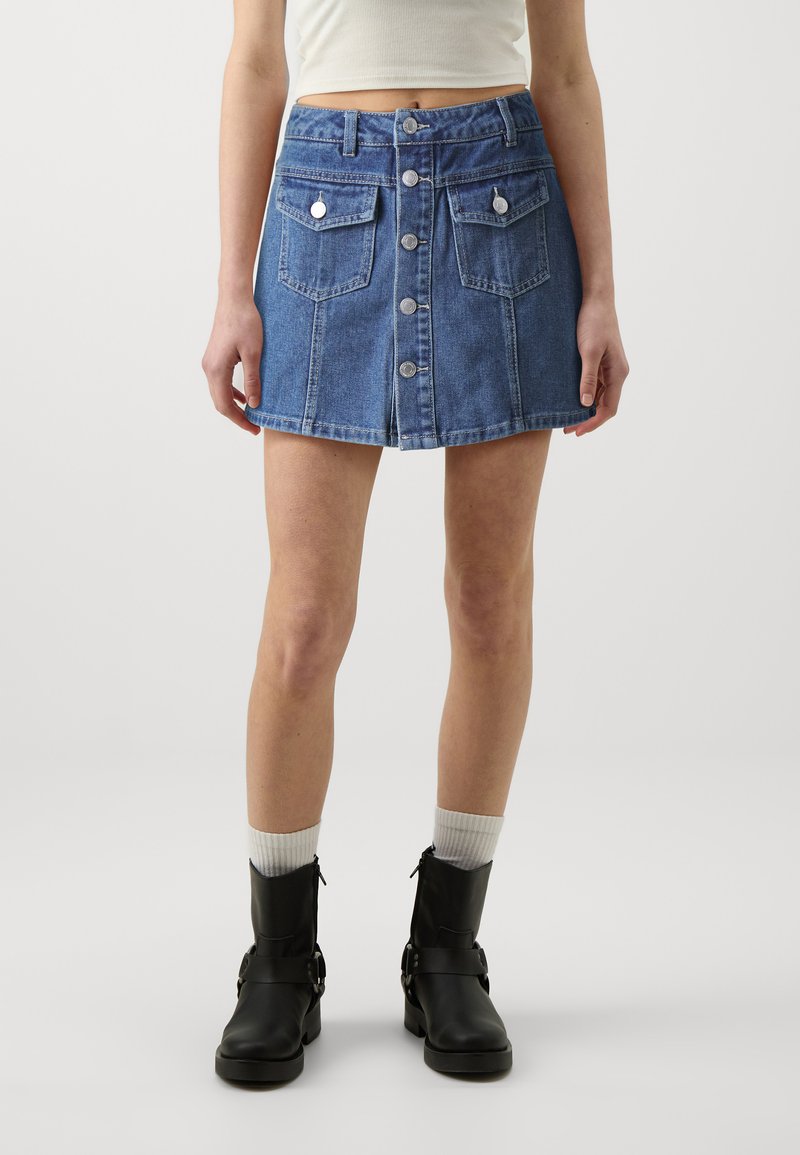 Denim mini suknja u plavoj boji s prednjim dugmetom, dva prednja džepa i ravnim rubom. Uparena s crnim čizmama do malih članka i bijelim čarapama.