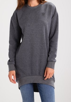 Femme portant un sweat-shirt long gris foncé avec des poignets et un ourlet côtelés, associé à un jean bleu, debout devant un fond uni.