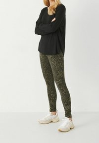 Haut noir à manches longues, leggings verts olive à motif à pois, et baskets blanches avec des accents beige clair, se tenant sur un fond clair.