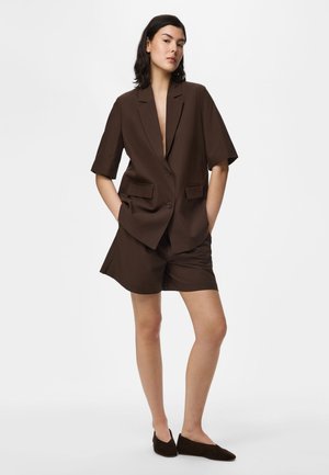 Femeie stând cu mâinile în buzunare, purtând un sacou scurt maro închis, pantaloni scurți asortați și pantofi slip-on închiși la culoare, pe fundal alb.