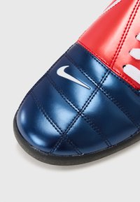 Red and navy soccer shoe with shiny, quilted texture; features prominent white Nike logo and black rubber outsole, laced design at the top.