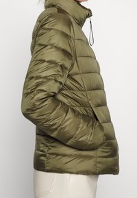Persona con una chaqueta acolchada de color verde oliva y cuello alto, con la mano en el bolsillo, combinada con pantalones de un color claro.
