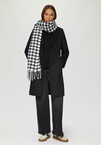Zwarte wollen jas, zwarte gebreide trui, grijze broek en een zwart-witte sjaal met houndstoothpatroon en franjes. Bruin-witte schoenen.