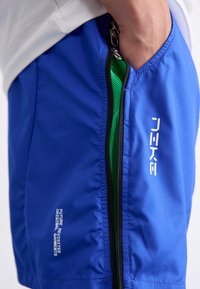 Blå sportsshorts lavet af letvægtsstof, med et grønt meshfor, sidelomme med lynlås og hvidt logo-tekst foran.