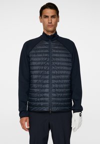 En man står självsäkert iklädd en mörk, isolerad jacka med dragkedja, som matchar med marinblå byxor och en vit golfhandske, mot en enhetlig bakgrund.