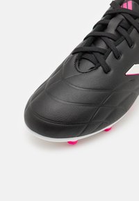 Botas de fútbol negras con parte superior de cuero sintético texturizado, que presentan un acento blanco y rosa, puntera redondeada y tacos rosas en contraste.