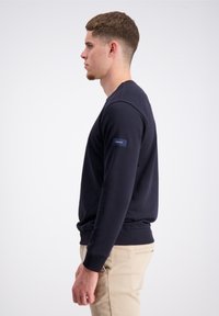 Granatowa bluza wykonana z miękkiego materiału, w fasonie crew neck, z prążkowanymi mankietami i dołem, z małą naszywką logo na lewym rękawie.