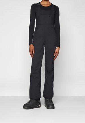 Person wearing black long-sleeve top and black zippered overalls with adjustable straps and black lace-up boots, standing against white background.