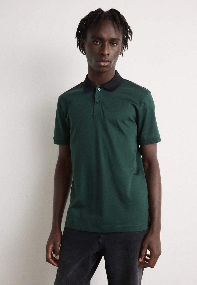 Zelená polokošeľa s čiernym golierom, krátkymi rukávmi a trojbotónovou légou, vystavená na modeli s neutrálnym pozadím.