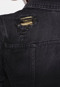 Svart jeansjacka med ett slitaget område på bakre axeln som avslöjar ett guldigt tyg under med fransiga kanter.