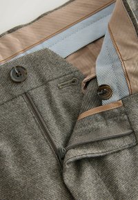 Brown textured fabric trousers with a zip closure, featuring dual brown buttons, a contrasting striped waistband, and a smooth inner lining.