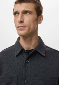 Camicia grigia scuro con chiusura a bottoni, colletto strutturato, due tasche frontali e bottoni neri. Texture liscia, design minimalista.