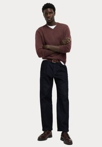 Pantalones de mezclilla azul oscuro combinados con un suéter marrón de cuello en V y zapatos marrones. El suéter tiene puños acanalados; el conjunto incluye un cinturón marrón.