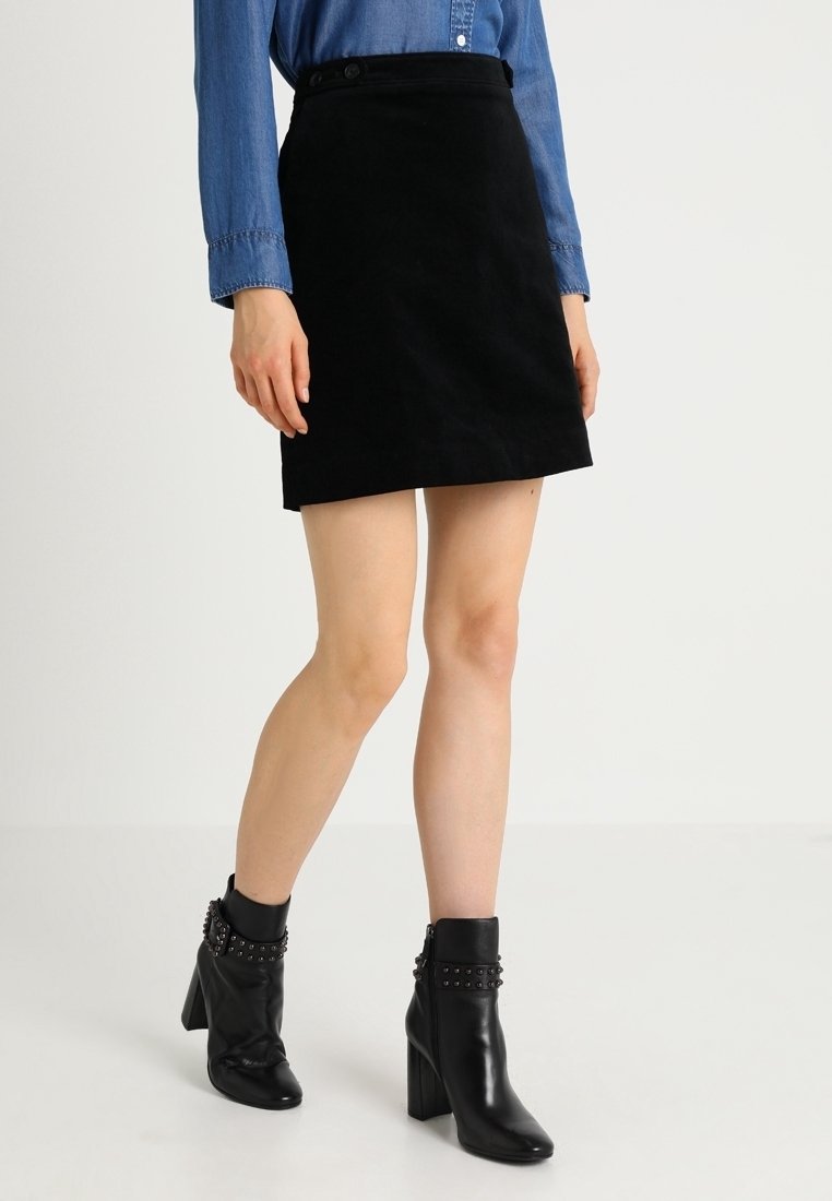 Femme portant une mini-jupe noire, une chemise bleue à manches longues boutonnée et des bottines noires à talons hauts avec des brides cloutées.