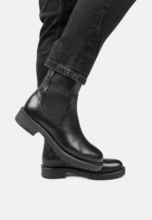 Sorte læder Chelsea-støvler med elastiske sidestykker båret med sorte jeans, den ene fod løftet over den anden mod en ensfarvet baggrund.