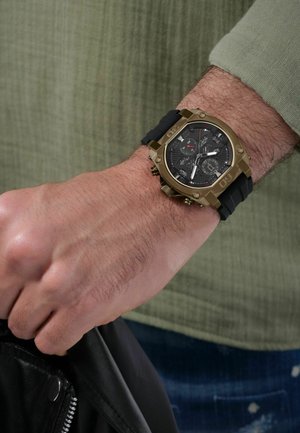 Black rubber strap watch with a bronze case, black dial featuring intricate details and red accents, worn on a hand grasping a jacket.