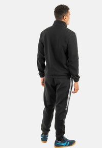 Zwarte trainingsoutfit met een jack met rits en elastische broek, voorzien van witte strepen aan de zijkanten en sportschoenen met blauwe accenten.