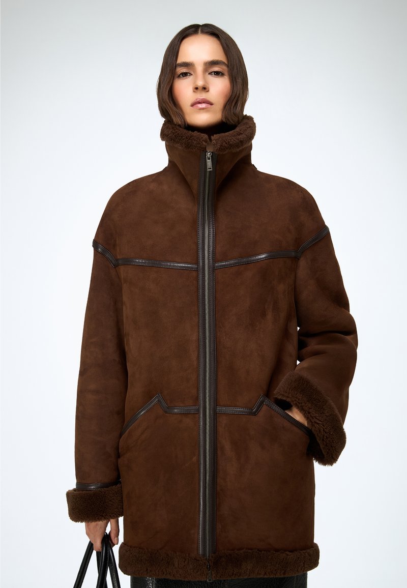 Bruine faux fur jas met een hoge kraag, ritssluiting aan de voorkant en lederen accenten. Voorzien van brede mouwen en een gestructureerde afwerking.