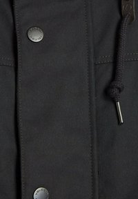 Dunkle Stoffjacke mit glatter Textur, auffälligen schwarzen Druckknöpfen und einer geflochtenen Kordel mit einem Metalldrehverschluss.