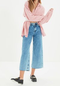 Nainen, joka pukeutunut vaaleanpunaiseen napilliseen paitaan, korkeavyötäröisiin leveälahkeisiin sinisiin farkkuihin ja mustiin loafereihin, joissa on kultainen solki, seisoo valkoista taustaa vasten.