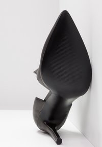 Chaussure à talon haut en cuir noir à bout pointu, posée sur le côté contre un mur d'angle blanc et le sol.