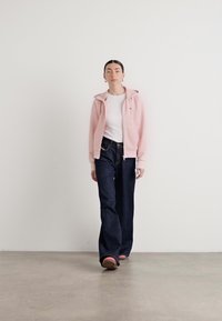 Sweat à capuche rose zippé sur un t-shirt blanc, associé à un jean large bleu foncé. Tissu lisse, coupe décontractée, minimalisme du branding sur le sweat.