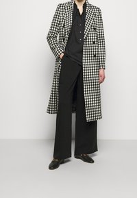 Lange zwart-wit geruite jas met een dubbelgeknoopt ontwerp, gecombineerd met een zwart shirt en wijde zwarte broek. Zwarte loafers.