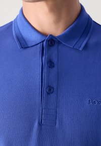 Gros plan d'une personne portant un polo bleu avec une patte de boutonnage à trois boutons et un logo brodé partiellement visible sur la poitrine.
