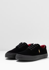 Mustat mokkakengät mustilla nauhoilla, joissa on kultainen logo sivulla ja punainen vuori, asetettu mustalle kumipohjalle.