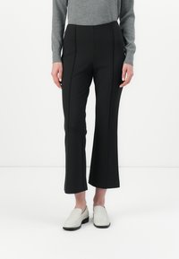 Pantaloni neri a palazzo con una superficie liscia, caratterizzati da dettagli di cucitura frontale, abbinati a scarpe slip-on di colore chiaro.