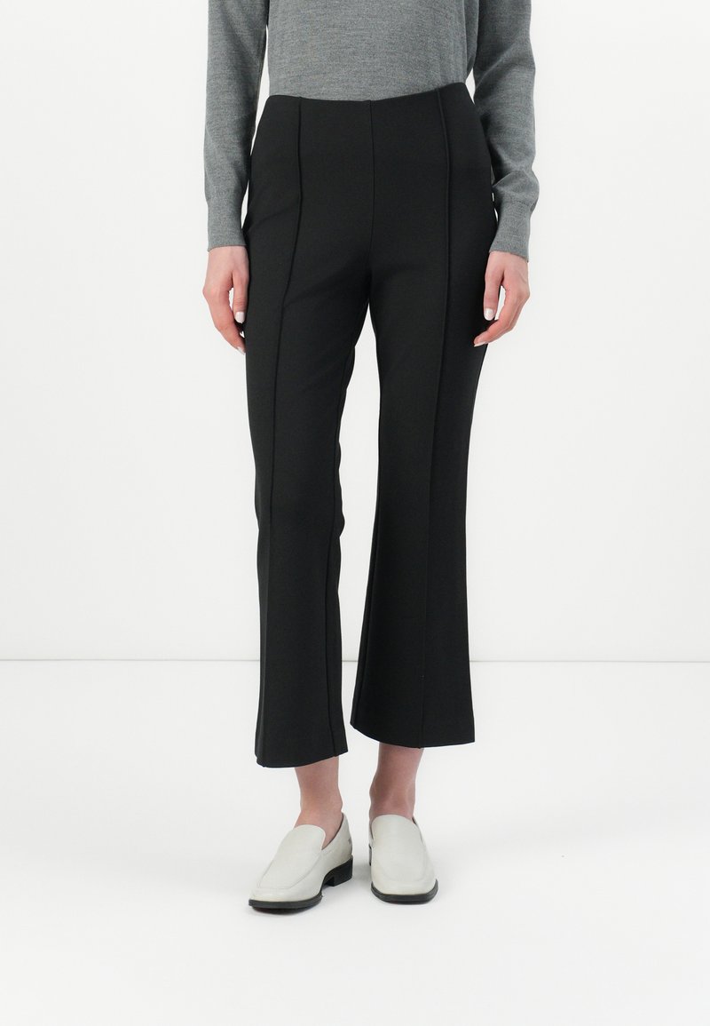 Pantaloni neri a palazzo con una superficie liscia, caratterizzati da dettagli di cucitura frontale, abbinati a scarpe slip-on di colore chiaro.