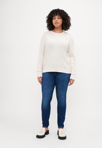Light cream knitted sweater with a textured pattern, paired with dark blue skinny jeans and off-white shoes with black soles.