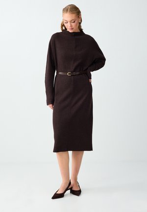 Kobieta w ciemnobrązowej, długorękawowej sukience midi z wąskim paskiem i dopasowanymi szpilkami w kolorze buta, stojąca na białym tle.