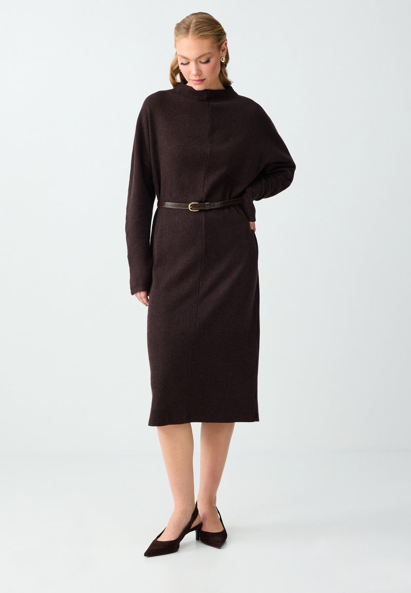 Femme portant une robe midi à manches longues de couleur marron foncé avec une ceinture fine et des escarpins pointus assortis, debout devant un fond blanc.