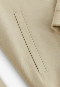 Beige tekstil med ribbet tekstur, der har en smal sidelomme, glatte sømme og en foldet manchet. Fokus på materiale og minimalistiske design detaljer.