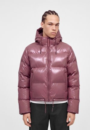 Joven con el pelo trenzado que lleva una chaqueta acolchada brillante de color malva con capucha, cremallera frontal y cordones, de pie frente a un fondo liso.
