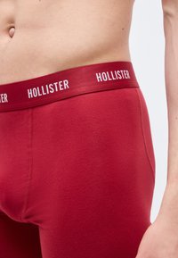 Κοντινή λήψη ενός ατόμου που φοράει κόκκινα εσώρουχα Hollister με λογότυπο στη μέση, μπροστά σε απλό φωτεινό φόντο.