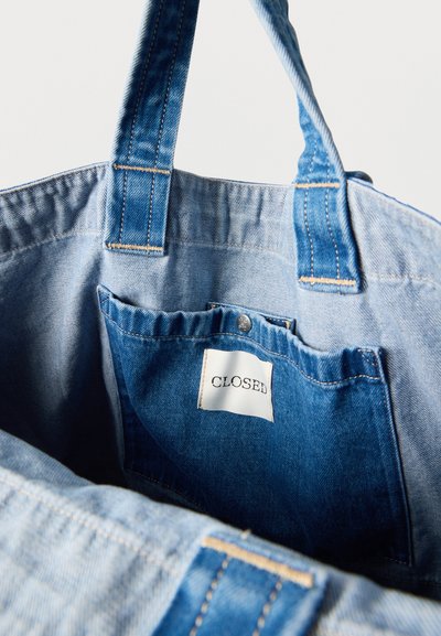 Τσάντα tote από ανοιχτό μπλε τζιν με πιο σκούρο μπλε εσωτερική τσέπη που διαθέτει λευκή ετικέτα με τη λέξη "CLOSED" και δύο φαρδιές λαβές.