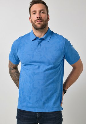 Mężczyzna z brodą i tatuażem, ubrany w niebieską koszulkę polo w wzory i ciemne dżinsy, stoi z rękami za plecami na jednolitym tle.