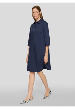 Frau, die in einem knielangen, dunkelblauen Hemdblusenkleid mit dreiviertel Ärmeln und silbernen, niedrigen Slingback-Schuhen vor einem schlichten grauen Hintergrund geht.