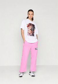 Hvit T-skjorte med grafisk trykk av sko, kombinert med rosa bukser med vide ben og sideliggende knapper, utstyrt med en liten svart etikett, brukt sammen med svarte og hvite joggesko.