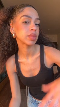 Fiatal nő hullámos copfban, aranykarika fülbevalóval, fekete ujjatlan felsőben és világoskék farmernadrágban, beltéren naplementekor.