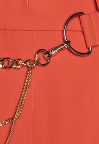 Tessuto corallo con una texture liscia. Presenta catene e accessori in metallo dorato, inclusi un anello e un attacco a clip.