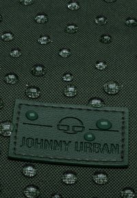 Zelená textúrovaná látka s vodnými kvapkami. Výrazná vyrazená značka s nápisom "JOHNNY URBAN" v tmavozelenej farbe. Zobrazuje vzorovaný povrch.