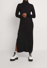 Schwarzes langärmliges Turtleneck-Kleid mit Seiten-Schlitzen; kombiniert mit weißen Schnürkampfstiefeln mit einer dicken schwarzen Sohle.