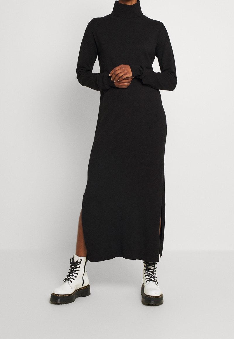 Schwarzes langärmliges Turtleneck-Kleid mit Seiten-Schlitzen; kombiniert mit weißen Schnürkampfstiefeln mit einer dicken schwarzen Sohle.