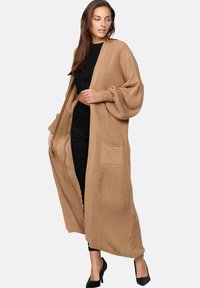 Lång, öppen cardigan i ljusbrunt stickat tyg. Har ribbad textur, vida ärmar och en framficka. Bärs över svarta kläder.